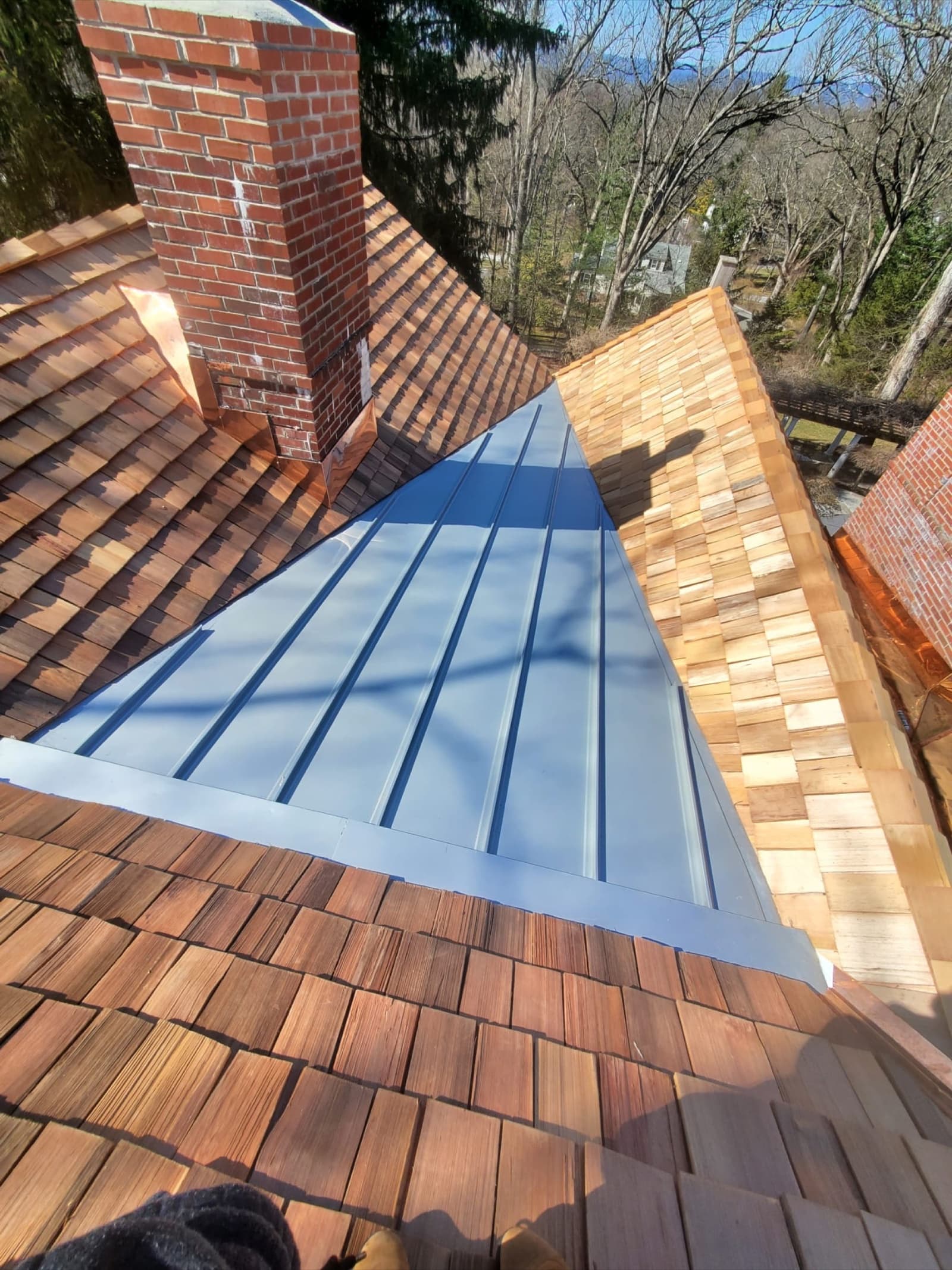 Standing-seam metal valley between cedar-shake slopes with a brick chimney by Zubar Roofing.